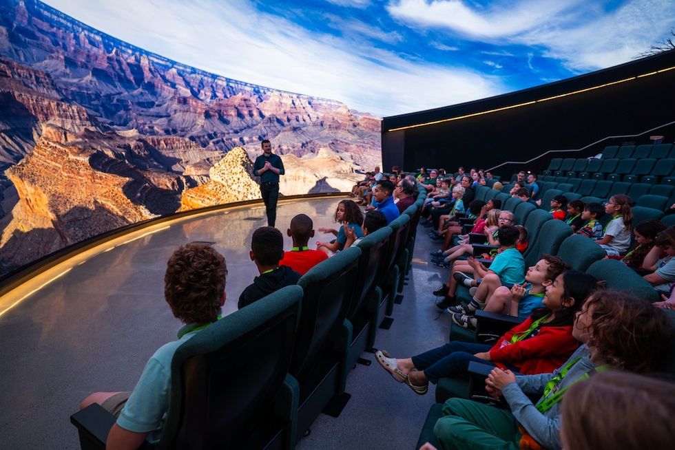 Audience watches a presenter in a theater with a panoramic Grand Canyon display.