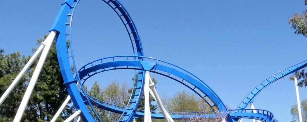 Blue roller coaster loops against a clear blue sky with trees in the background.
