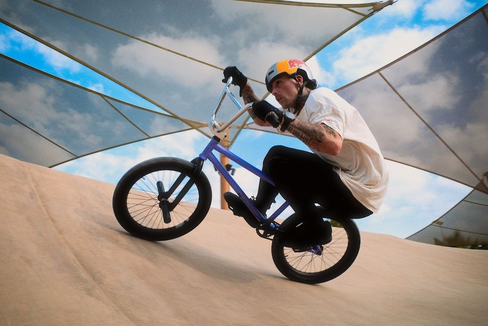 BMX rider with tattoos and helmet performing on a sunny, canopy-covered ramp at Adrena