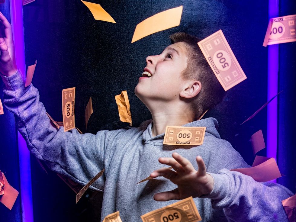 Boy in gray hoodie catching Monopoly money in a booth with purple lighting.