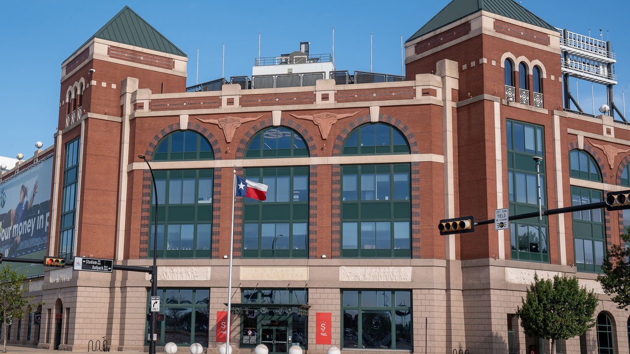 Brick building with arched windows and a Texas flag in front, under a clear blue sky.