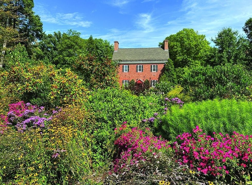 Brick house at Mt. Cuba Center, surrounded by vibrant, blooming garden and green foliage under a blue sky.
