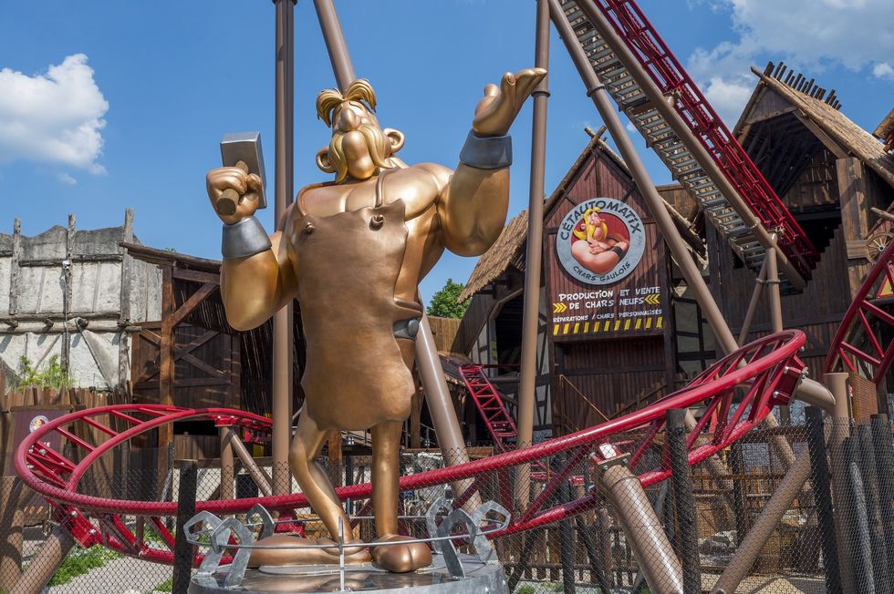 Bronze cartoon statue with a hammer, red roller coaster, wooden buildings, and blue sky - Ce\u0301tautomatix coaster at Parc Asterix