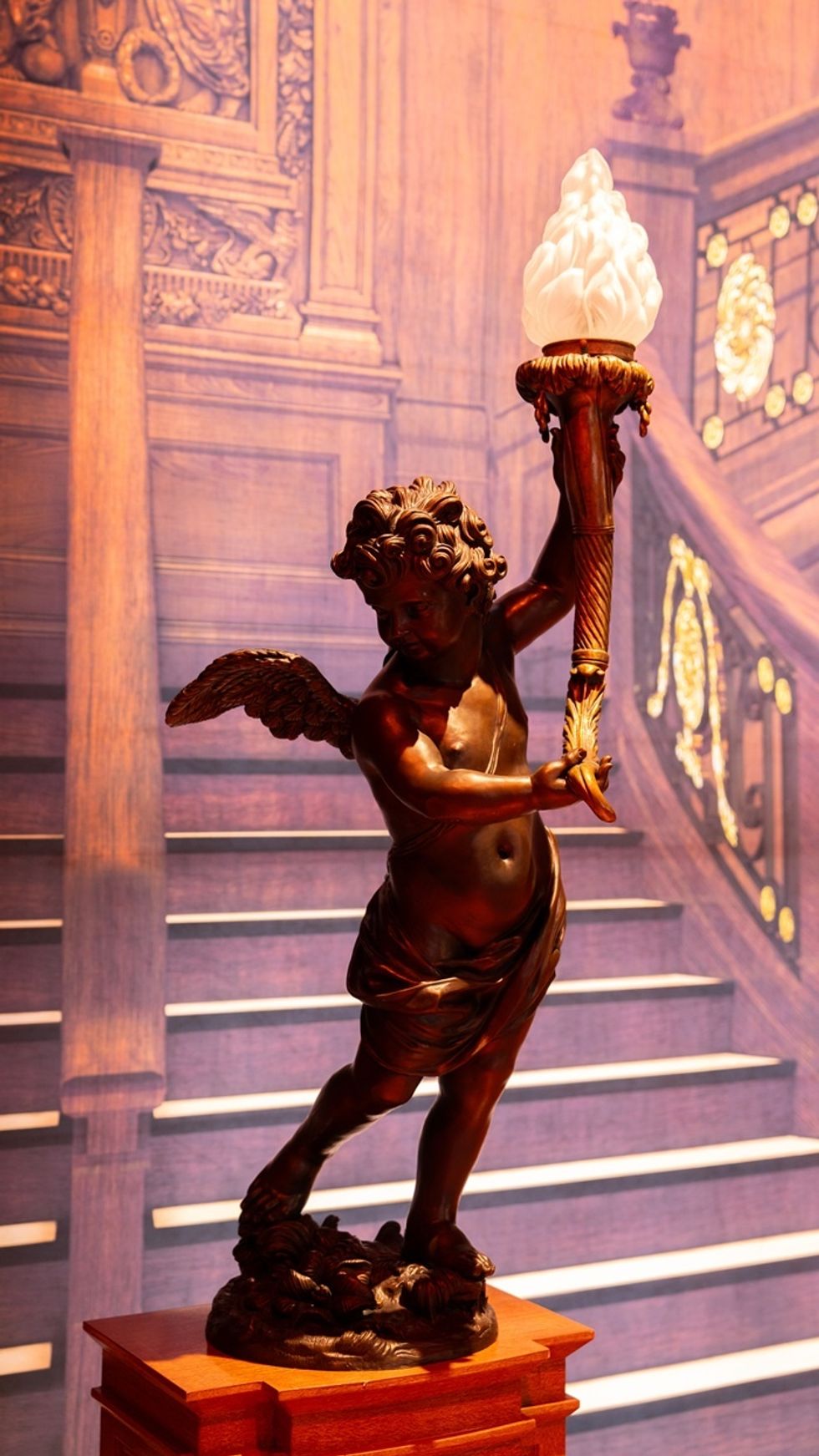 Bronze cherub statue holding a torch, set against an ornate wooden staircase backdrop in Titanic: An Immersive Voyage.