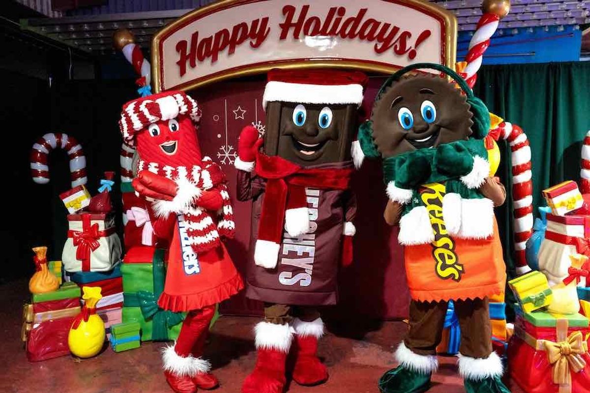 Candy mascots in holiday attire in front of a "Happy Holidays" sign with gifts.
