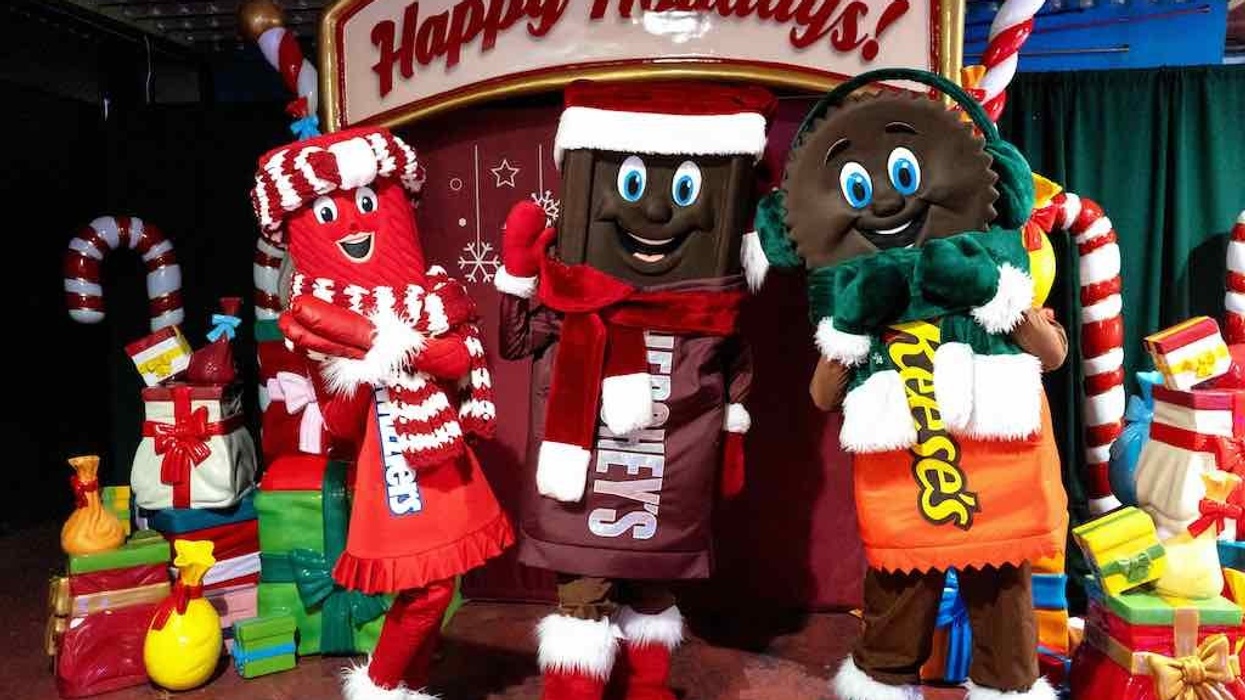 Candy mascots in holiday attire in front of a "Happy Holidays" sign with gifts.