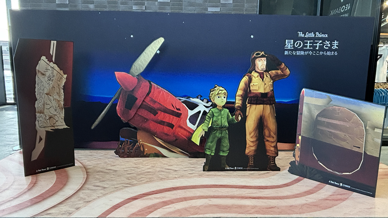 Cardboard cutouts of "The Little Prince" and pilot beside a red plane at a display.