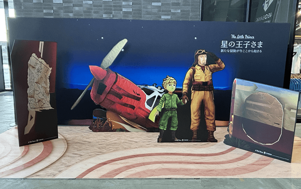 Cardboard cutouts of "The Little Prince" and pilot beside a red plane at a display.