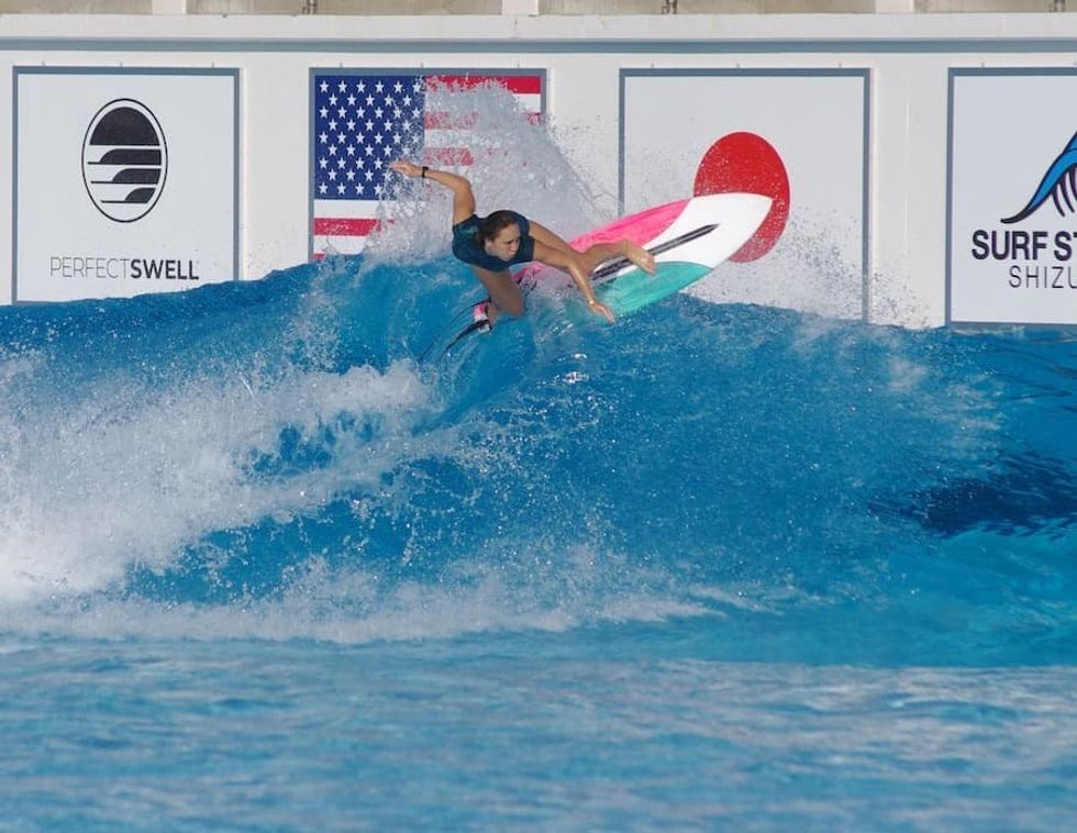Carissa Moore on a colorful board riding a wave at an indoor surfing facility in Japan with flags in the background.