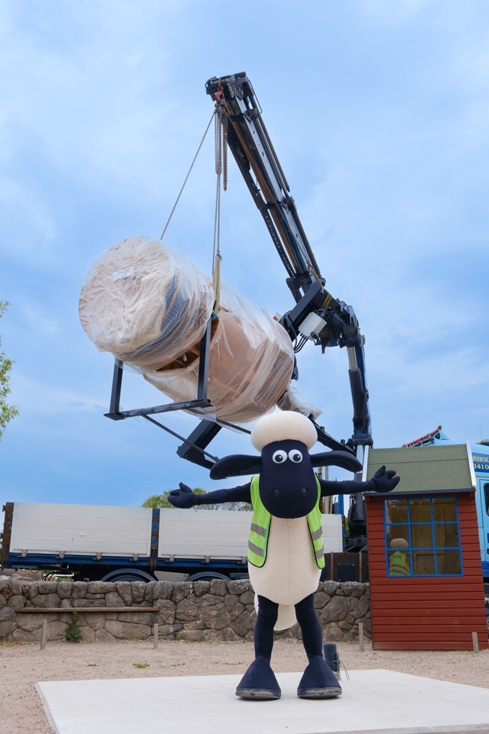 Cartoon sheep mascot, Shaun the Sheep, with a vest in front of a crane lifting a wrapped object, blue sky above.