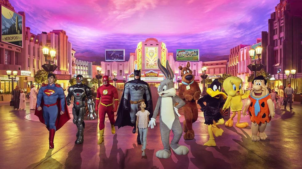 Characters and a child walking in Warner Bros. park with a vibrant cityscape background.
