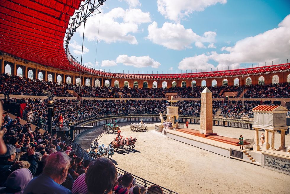 Chariot race in a crowded Roman-style arena under a bright blue sky at Puy du Fou