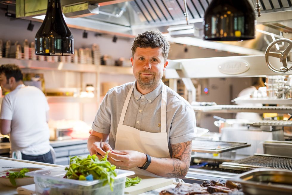 Chef Robin Gill in a bright kitchen preparing fresh ingredients, wearing a white apron.