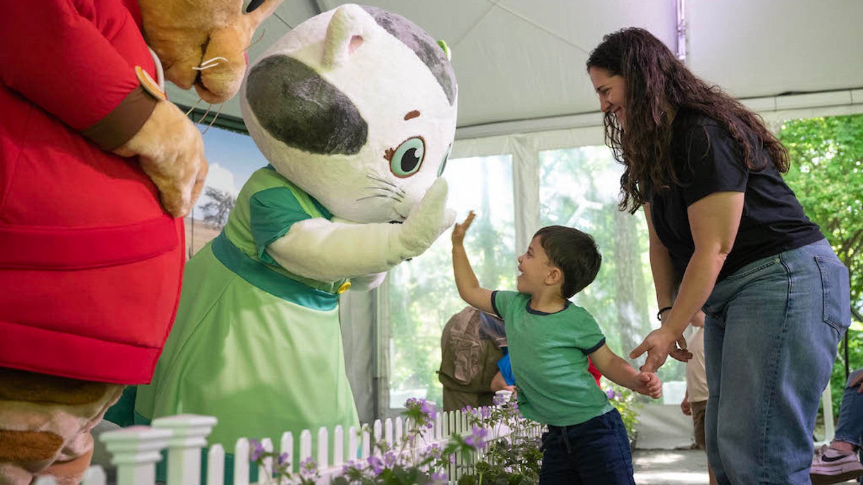 Child high-fives a costumed character, held by a smiling adult in a tent setting.