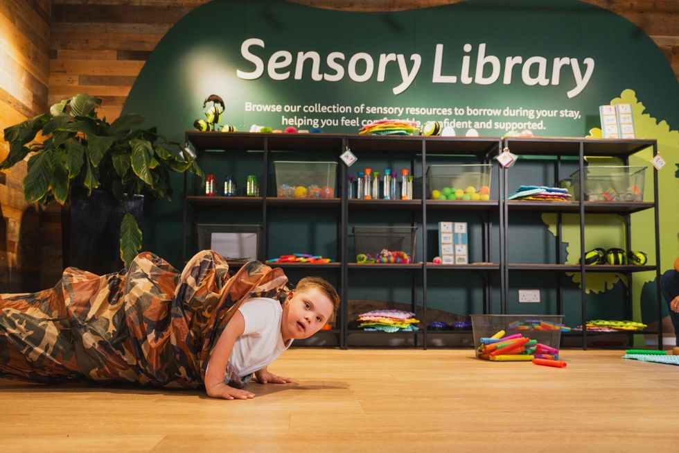 Child in a camouflage sack at a sensory library with shelves of colorful items.