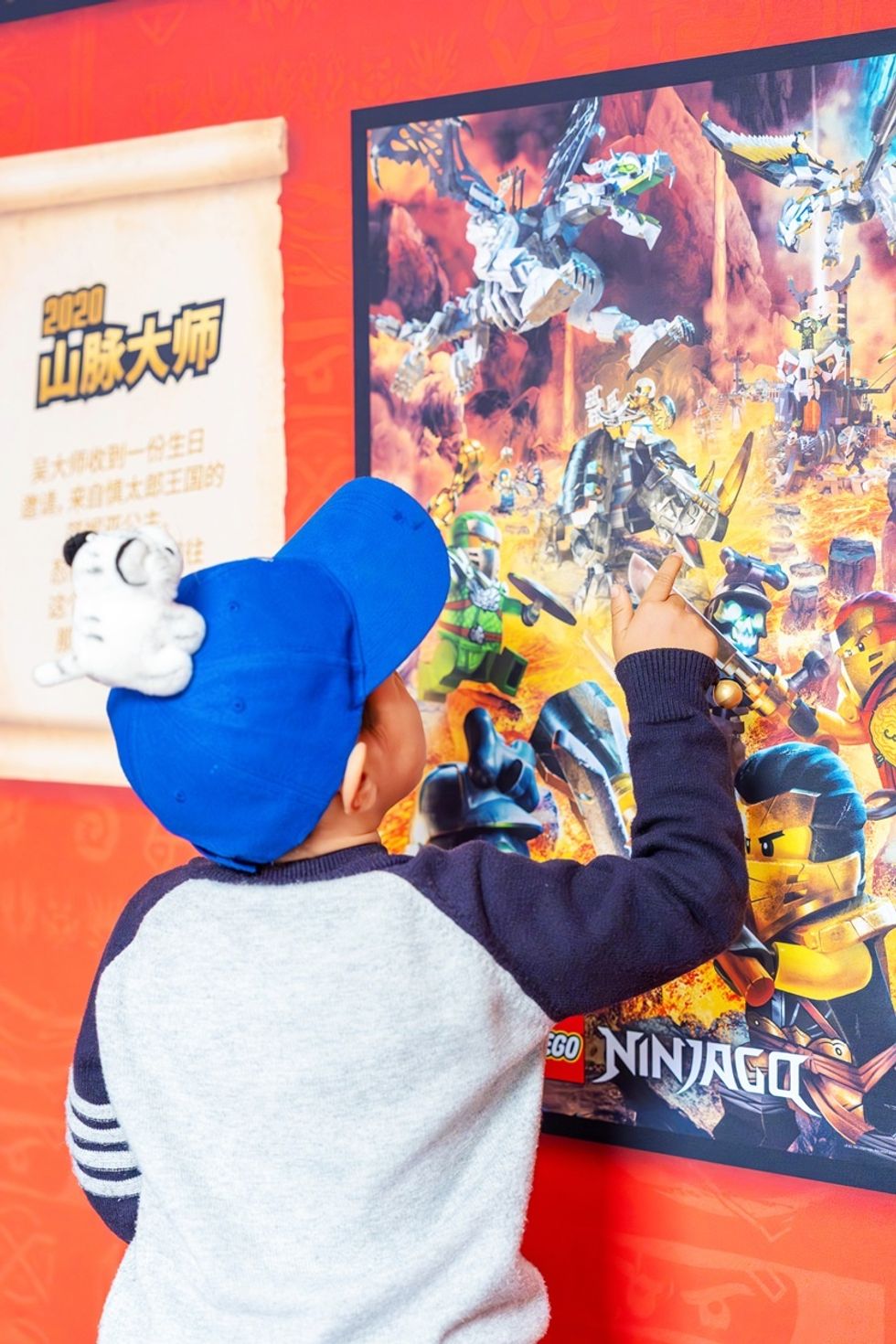 Child in blue cap points at colorful LEGO Ninjago poster on red wall.