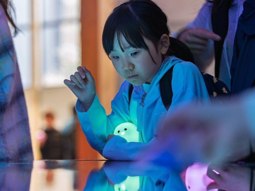 Child in blue jacket holds glowing toy, intently watching colorful table display.