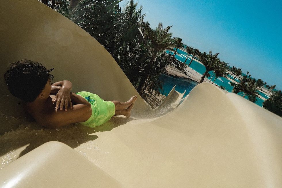 Child in green trunks sliding down a waterslide at Adrena with palm trees and pool in the background.