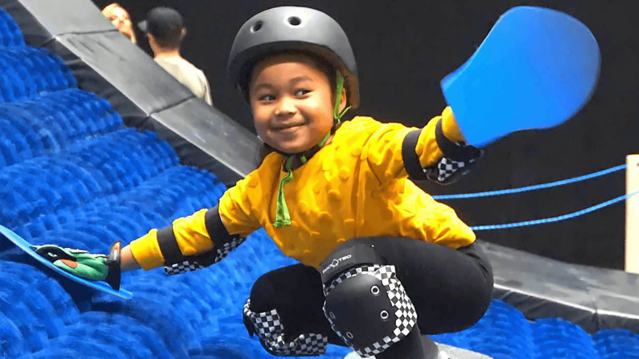 Child in helmet and pads snowboards indoors on a blue ramp.