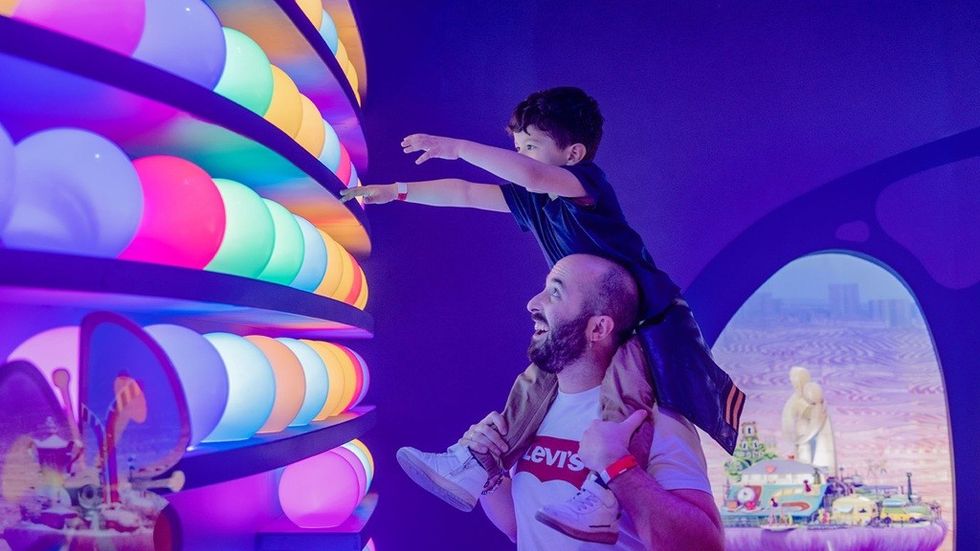 Child on adult's shoulders reaching for colorful glowing wall in a playful, immersive setting.