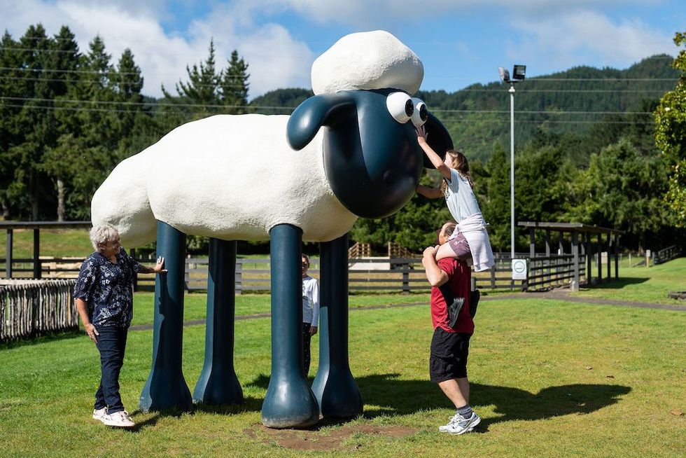Child on shoulders touches giant Shaun the Sheep statue's eye, with an adult nearby, in a grassy park.