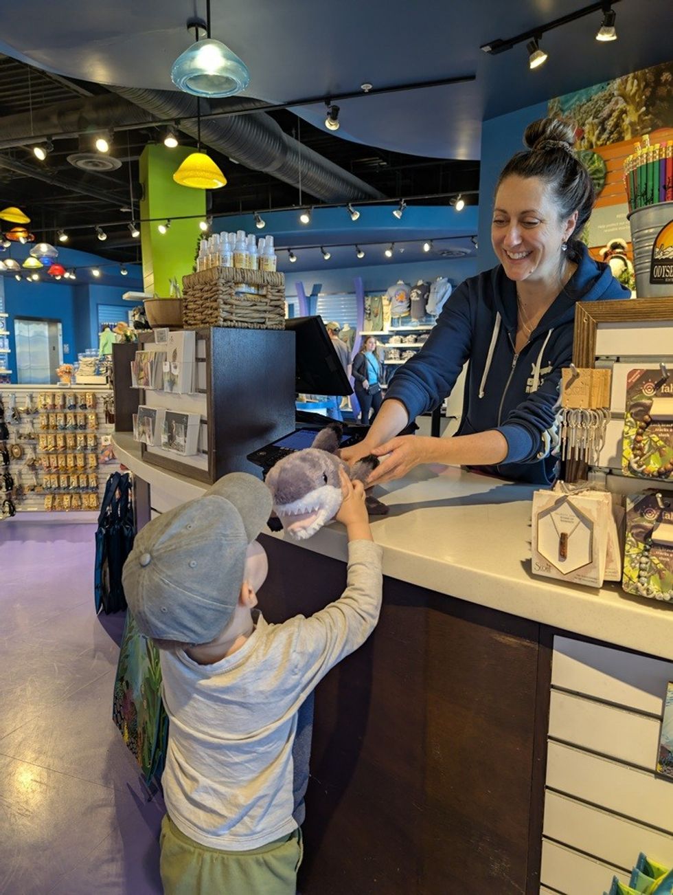Child receiving plush shark from shopkeeper in a colorful souvenir store.