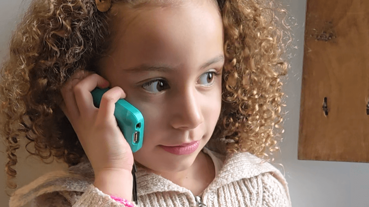 Child with curly hair holds a turquoise phone to her ear, wearing a knitted sweater.