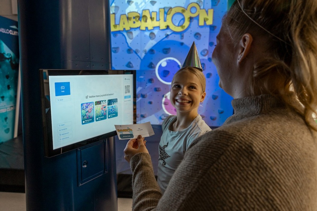 Child with party hat smiles at event kiosk, colorful climbing wall from Valo Motion in background.