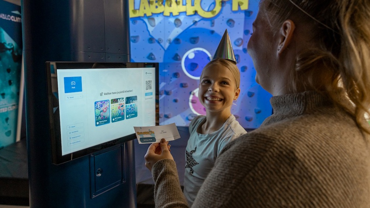 Child with party hat smiles at event kiosk, colorful climbing wall from Valo Motion in background.