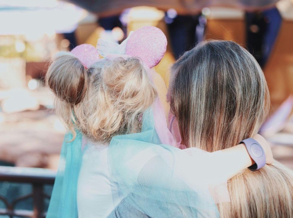 Child with pink Mickey ears and adult embracing, facing away outdoors.