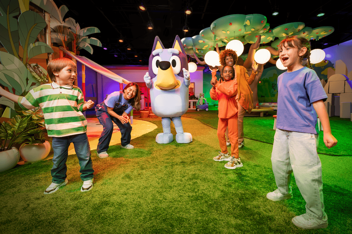Children and adults playing with a large, blue cartoon character in a colorful indoor playground.