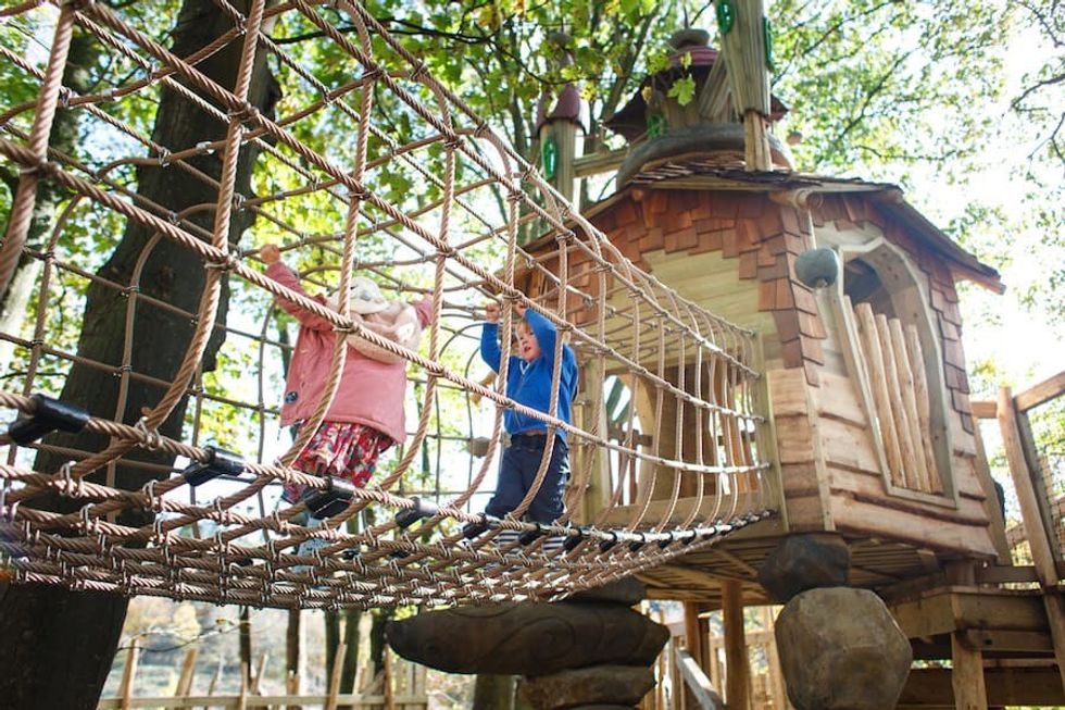 Children crossing a rope bridge to a treehouse in a wooded playground.