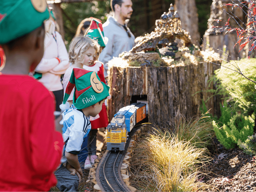 Children in elf hats watch a model train emerging from a tunnel in a garden setting.