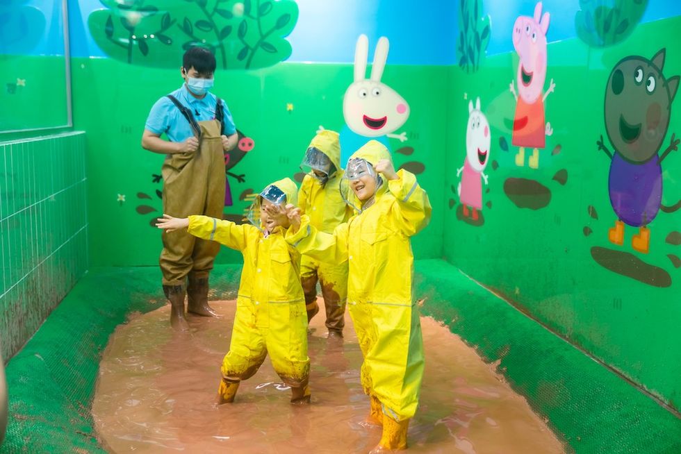 Children in yellow rain gear play in a muddy area with cartoon animal decorations.