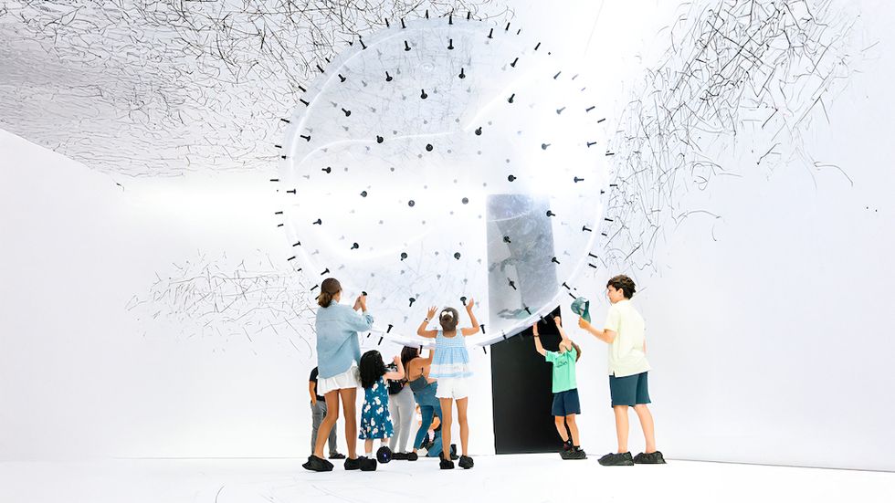 Children interact with a large, transparent ball in a white room filled with fine lines.