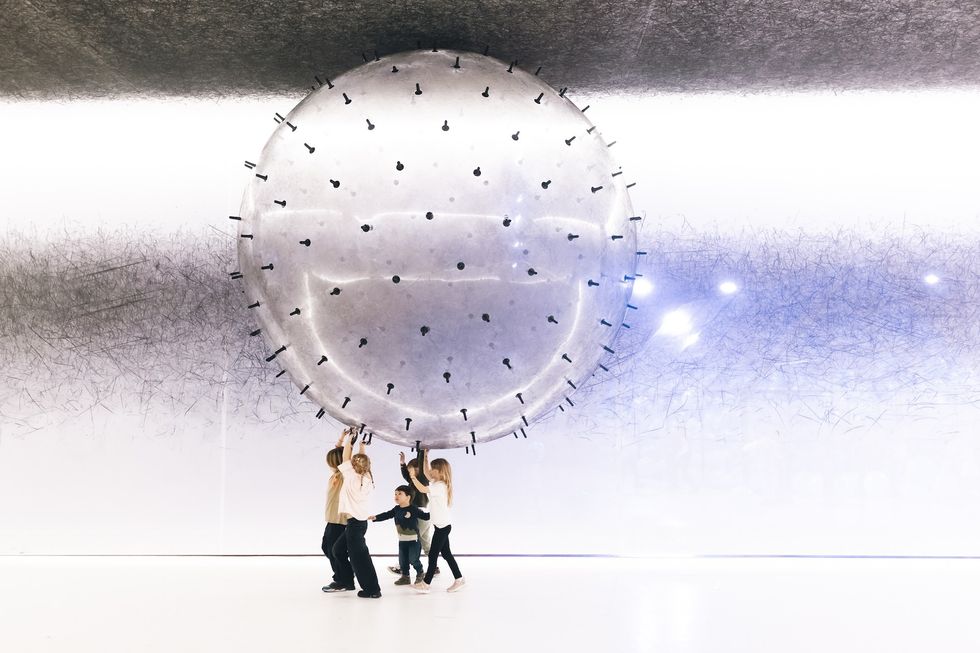 Children lift a large, spiky, transparent ball indoors against a textured wall.