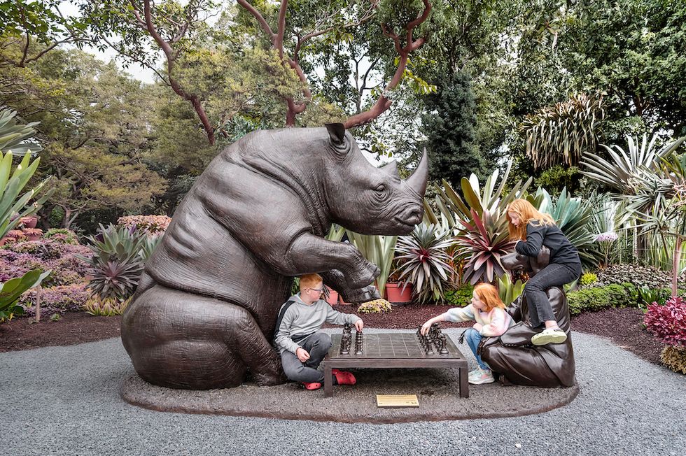 Children play chess with a large rhino statue in a garden setting.