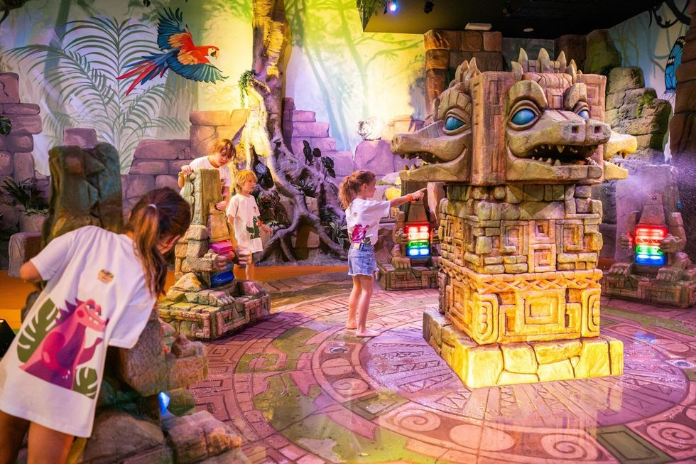 Children play in a colorful, jungle-themed indoor adventure area with stone structures.