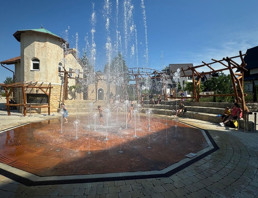 Children playing in a plaza fountain on a sunny day, surrounded by rustic architecture.
