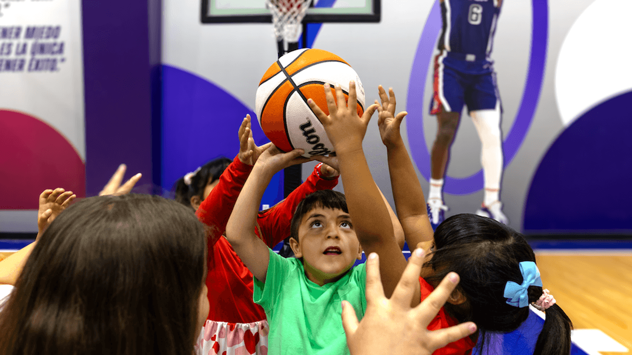 Children reach for a basketball on an indoor court under a mural.
