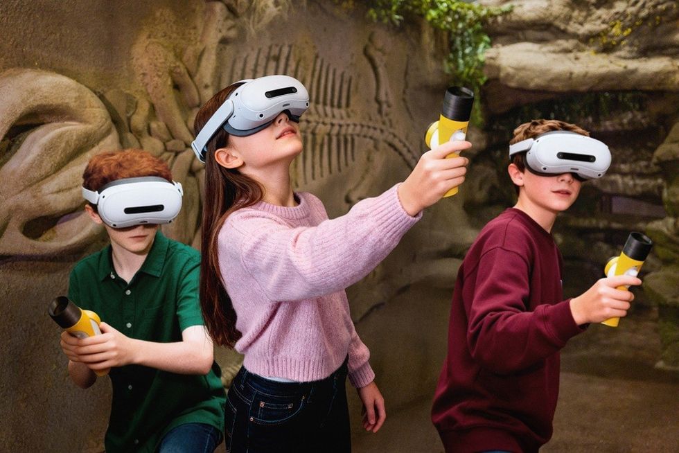 Children with VR headsets and controllers explore a virtual prehistoric environment.