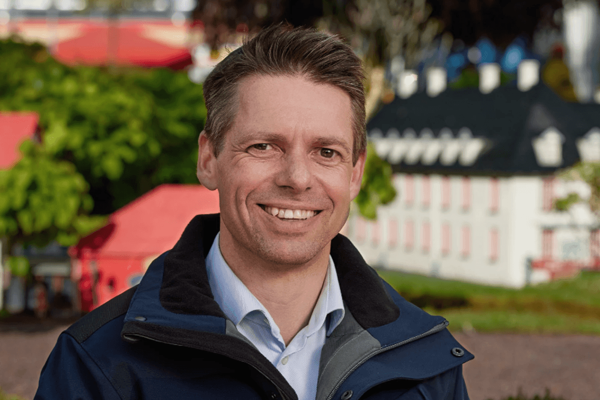 ​Christian Woller smiling outside, wearing a blue jacket, with colorful buildings in the blurred background.