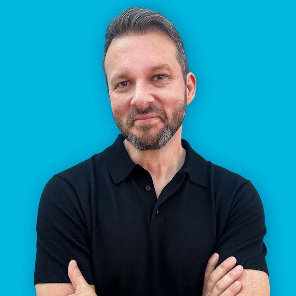 Chrsi Grap, Man in a black shirt smiling with arms crossed against a blue background.