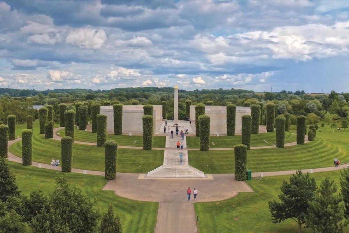 Circular memorial landscape with tall hedges, central walkway, and distant cloudy sky.