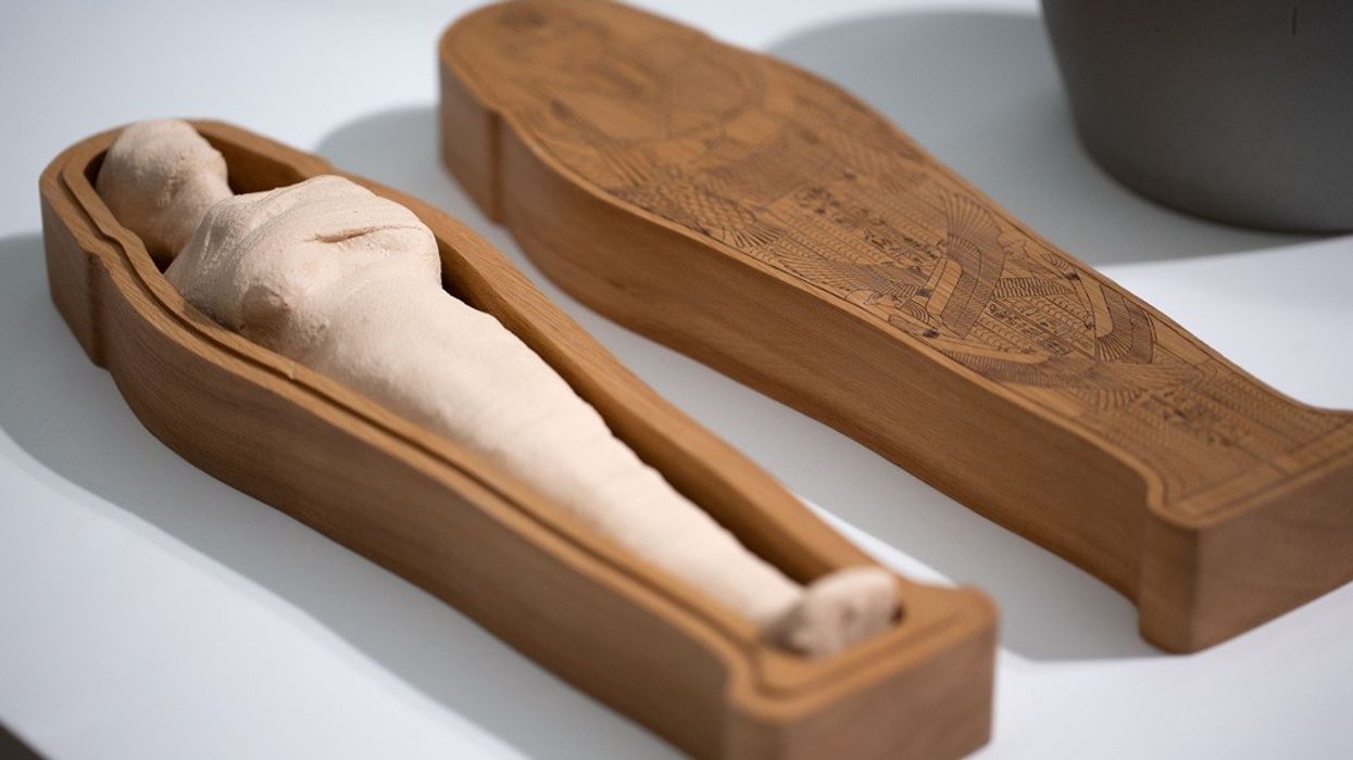 Clay mummy in a wooden sarcophagus with hieroglyphic engravings, on a white surface.