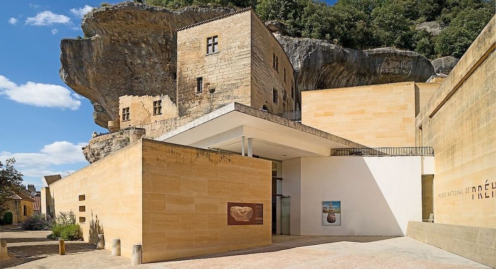 Cliffside museum with stone walls under a rocky overhang, surrounded by greenery.