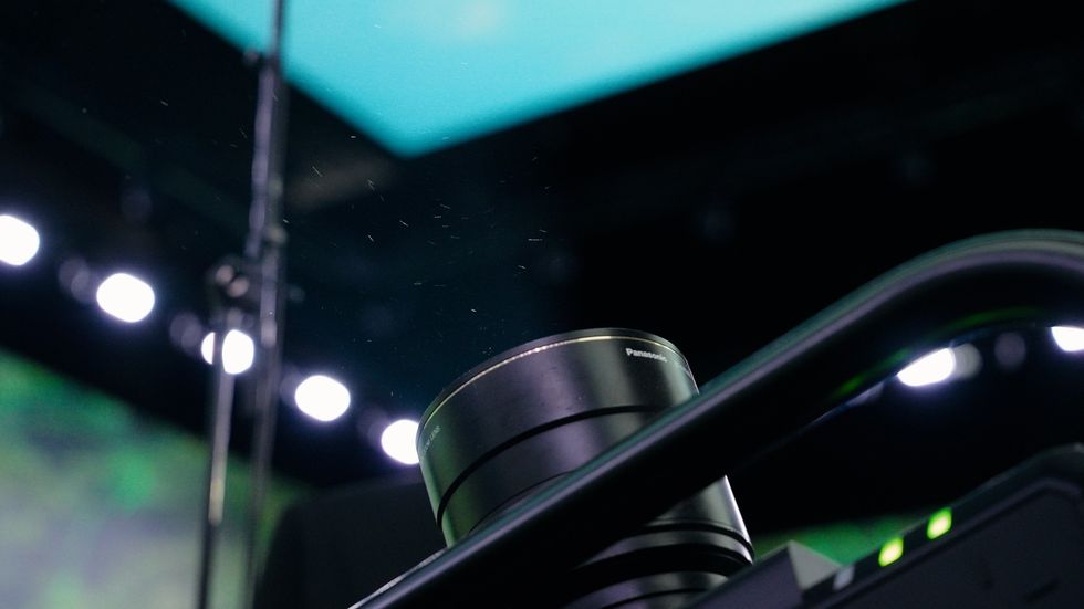 Close-up of a camera lens with blurred stage lights and microphone in the background.