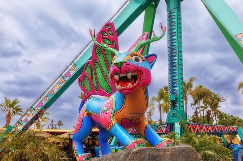 Colorful alebrije sculpture with antlers in a theme park setting, cloudy sky background.