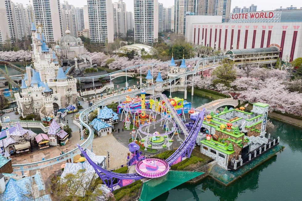 Colorful amusement park with castle, rides, and cherry blossoms, city skyline in background.