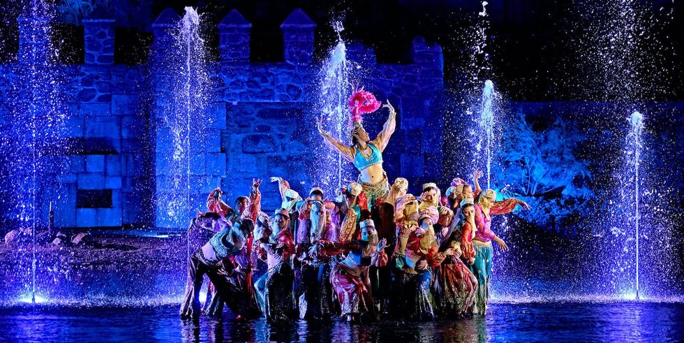 Colorful dance performance with fountains and blue-lit stone wall background.